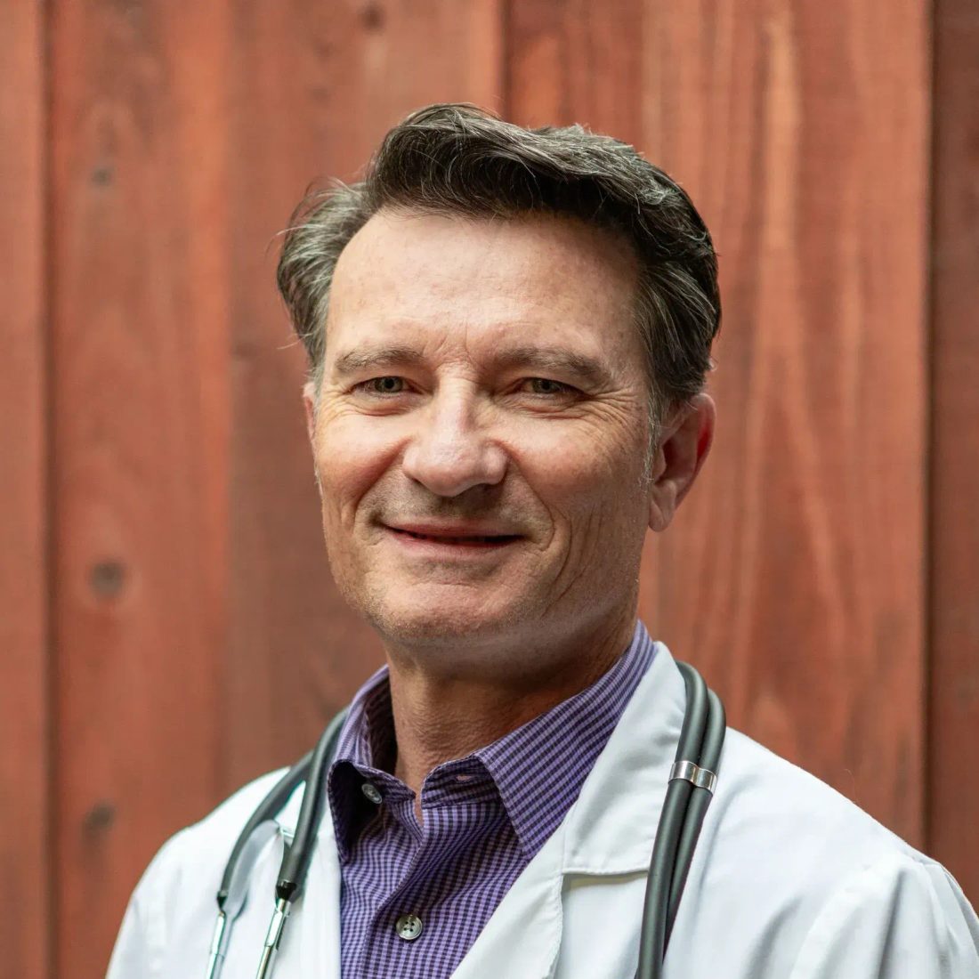 Smiling male veterinarian in white coat with stethoscope against wood wall